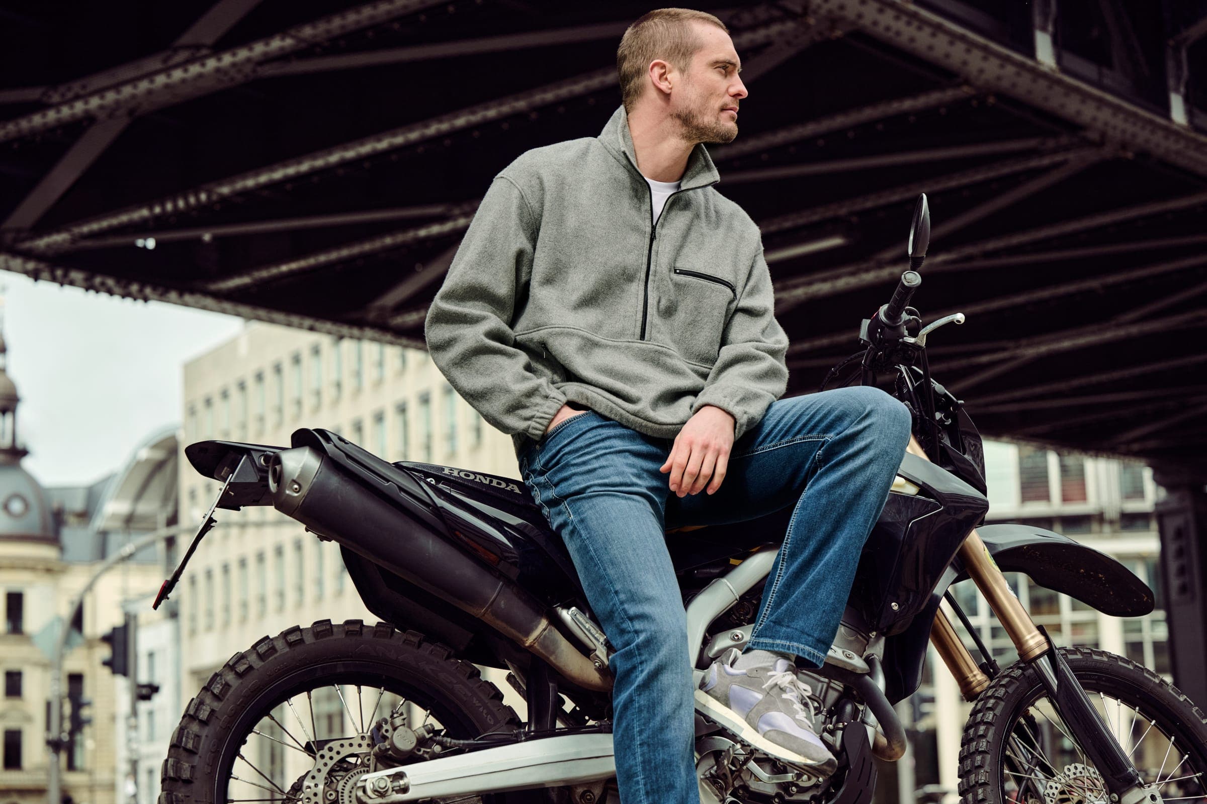 Man met MAC-spijkerbroek, zittend op een motorfiets onder een stalen brug